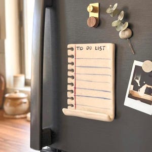 Könnte beinhalten: Ein Keramik-Notizblockmagnet in Form eines linierten Notizbuchblatts mit Spiralbindung. Die cremefarbene Oberfläche zeigt den Text TO DO LIST in schwarzer Tinte, blaue horizontale Linien, eine rote vertikale Randlinie und eine kleine Ablage.