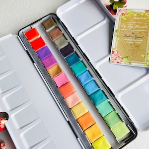 Pode incluir: Uma paleta de aquarela de metal branco com uma variedade de quadrados de tinta coloridos. A paleta contém uma variedade de tons, incluindo vermelho, roxo, rosa, laranja, amarelo, verde e azul. Um pequeno livro com detalhes florais também é visível.