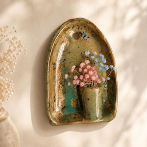 Peut inclure: Un vase mural en céramique avec une plaque arrière arquée présentant un vernis tacheté vert et brun. Une petite poche cylindrique contient un bouquet de fleurs séchées gypsophile dans des tons rose clair et bleu doux. Un trou est en haut.