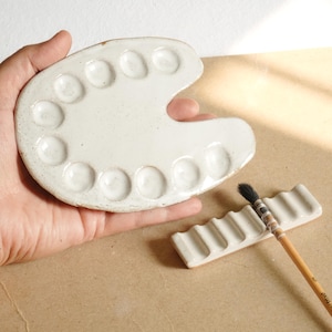 Puede incluir: Paleta de artista de cerámica blanca con múltiples pozos de pintura, sostenida en una mano. También hay un soporte para pinceles a juego y un pincel. La paleta y el soporte para pinceles están sobre una superficie marrón.