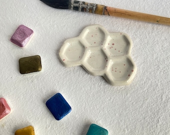 May include: A white ceramic paint palette with six hexagonal wells, each with a speckled surface. The palette is surrounded by small squares of different colored paint.