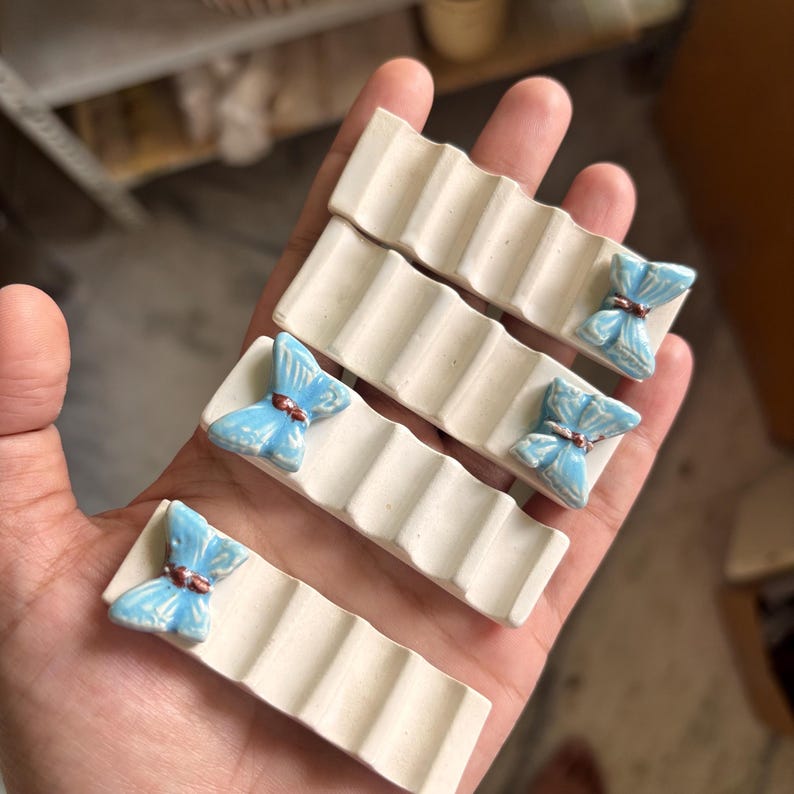 Puede incluir: Una mano sostiene cuatro jaboneras rectangulares de cer&aacute;mica blanquecina. Cada plato tiene un dise&ntilde;o ondulado y un acento de mariposa azul claro con un centro marr&oacute;n. Los acentos de mariposa est&aacute;n en el extremo de cada plato.