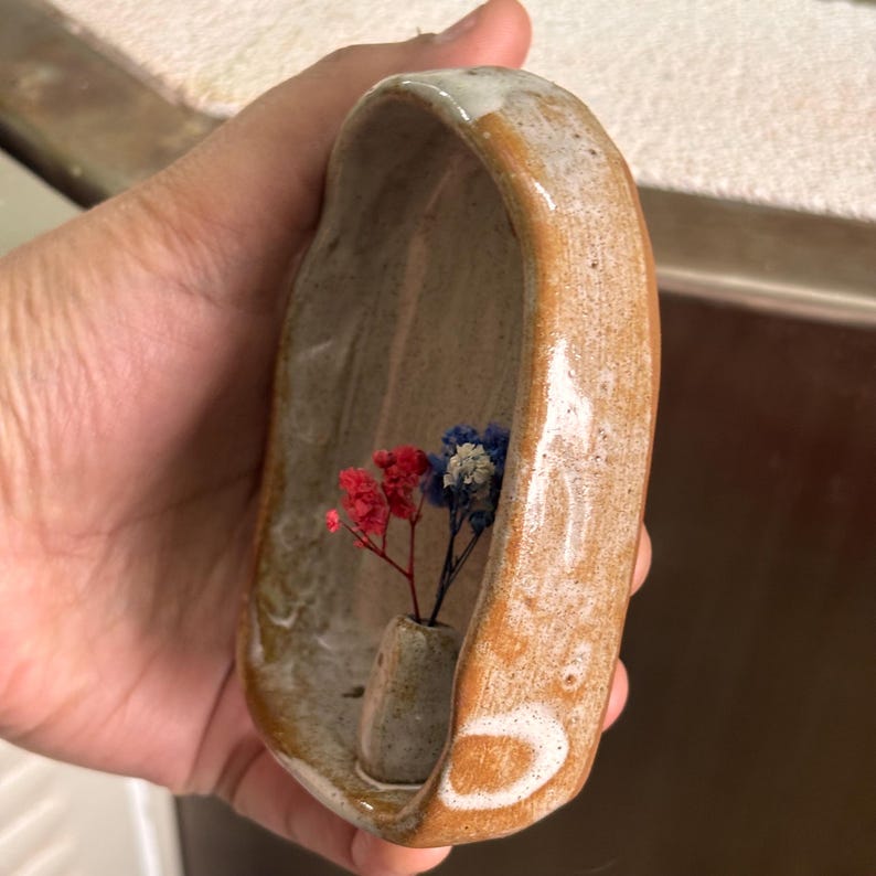 May include: A handmade ceramic vase with a curved, oblong shape. The vase is a warm brown color with white accents and contains a small arrangement of red, white, and blue dried flowers. The vase is designed to hold small floral arrangements.