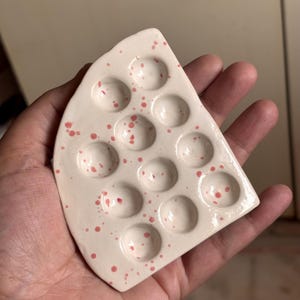 Puede incluir: Una paleta de pintura de cerámica con forma de cuña de queso. La paleta es de color blanquecino con motas rosas y tiene 12 pozos circulares. La paleta se sostiene en una mano.