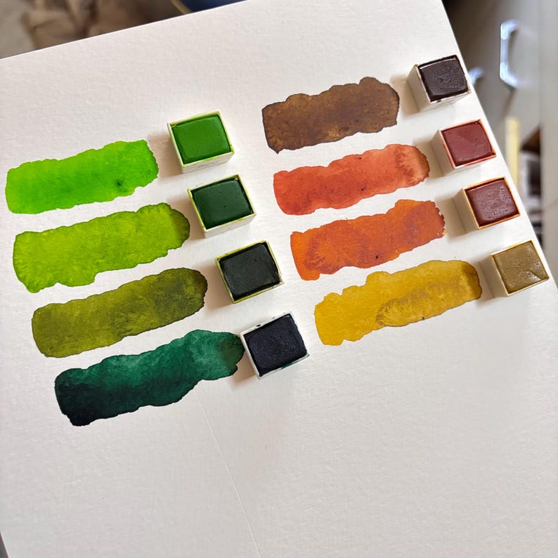 May include: A collection of watercolor paints in various shades of green, brown, orange, and yellow. Each color is displayed as a swatch and in a small square pan. The swatches are on a white paper surface.