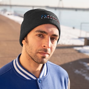 May include: A black knit beanie with the word "FREEDOM" embroidered in red, white, and blue. The beanie is worn by a person with a blue jacket.