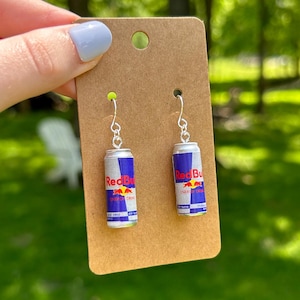 Puede incluir: Un par de pendientes colgantes con dijes en miniatura de latas de bebida energ&eacute;tica Red Bull en colores plata, azul y rojo. Los dijes est&aacute;n sujetos a ganchos de metal plateado y se presentan sobre una tarjeta de cart&oacute;n marr&oacute;n para joyer&iacute;a.