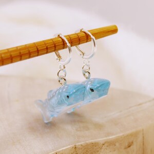 May include: A pair of silver-toned hoop earrings featuring translucent light blue whale shark charms. The earrings are suspended from a wooden chopstick, showcasing the detailed molded texture of the shark figures with visible dark eyes.