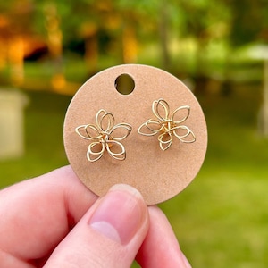 May include: A pair of gold-colored wire stud earrings shaped like five-petaled flowers. The earrings are attached to a circular brown cardboard display card with a small hole at the top for hanging, showcasing the intricate looped wire design.