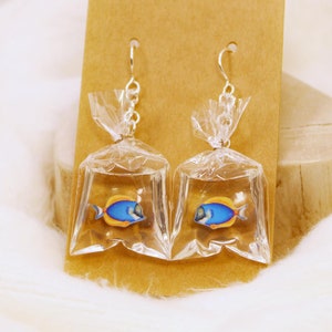 May include: A pair of dangle earrings featuring a blue and yellow fish inside a clear plastic bag. The earrings are hanging from a brown card.