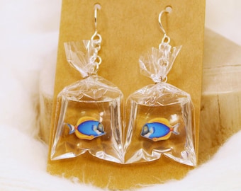 May include: A pair of dangle earrings featuring miniature clear plastic bags filled with water and a small blue and yellow tang fish graphic. The earrings are attached to silver-toned metal hooks and are displayed on a rectangular brown card.