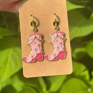 May include: A pair of gold-toned hoop earrings with pink and white cowboy boot charms. The boots have a star pattern and a darker pink trim. The earrings are displayed on a brown card against a backdrop of green foliage.