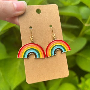 May include: A pair of rainbow-shaped earrings with red, orange, yellow, green, blue, and black enamel. The earrings are attached to a brown cardboard earring display card with a gold-colored hook.