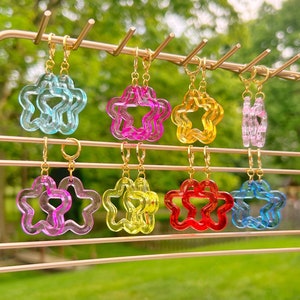 May include: Eight pairs of translucent, star-shaped earrings hanging on a gold metal display rack. The earrings come in various colors including light blue, bright pink, yellow, light purple, red, and dark blue, all featuring gold-toned hoops.