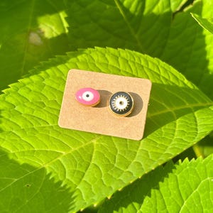 May include: Two enamel eye stud earrings. One is pink with a white iris and black pupil, the other is black with a white iris and black pupil. Both have gold-colored metal borders. The earrings are on a small, brown, rectangular card.
