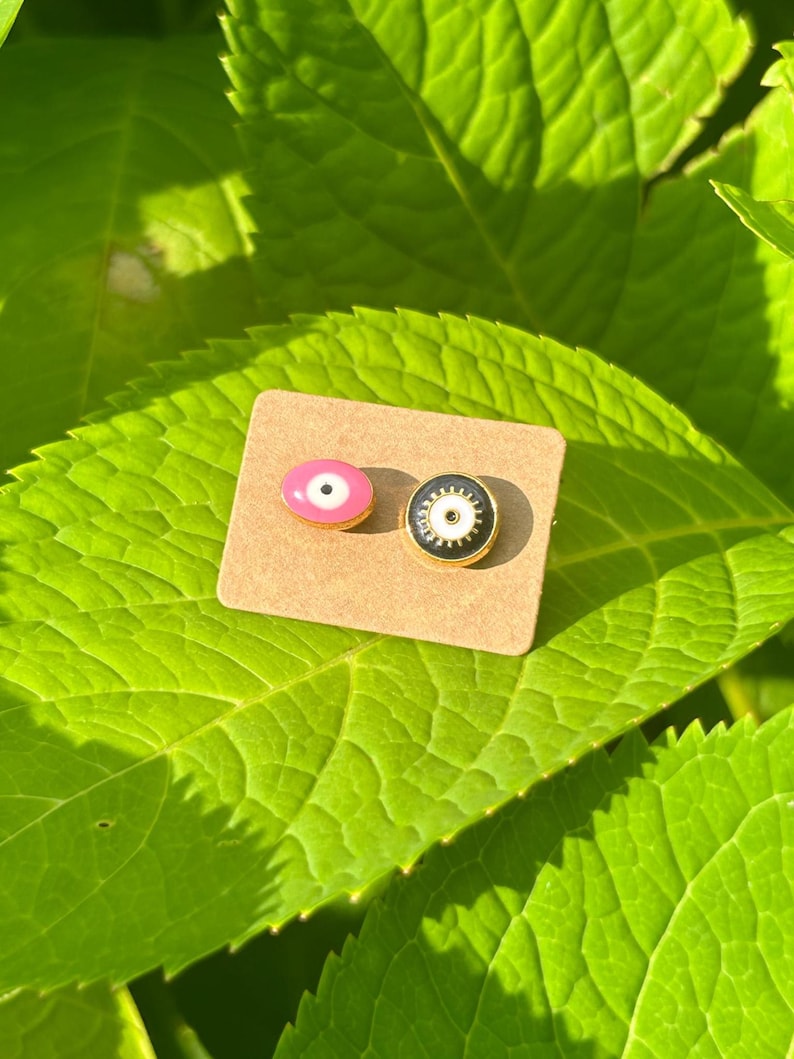 May include: Two enamel eye stud earrings. One is pink with a white iris and black pupil, the other is black with a white iris and black pupil. Both have gold-colored metal borders. The earrings are on a small, brown, rectangular card.