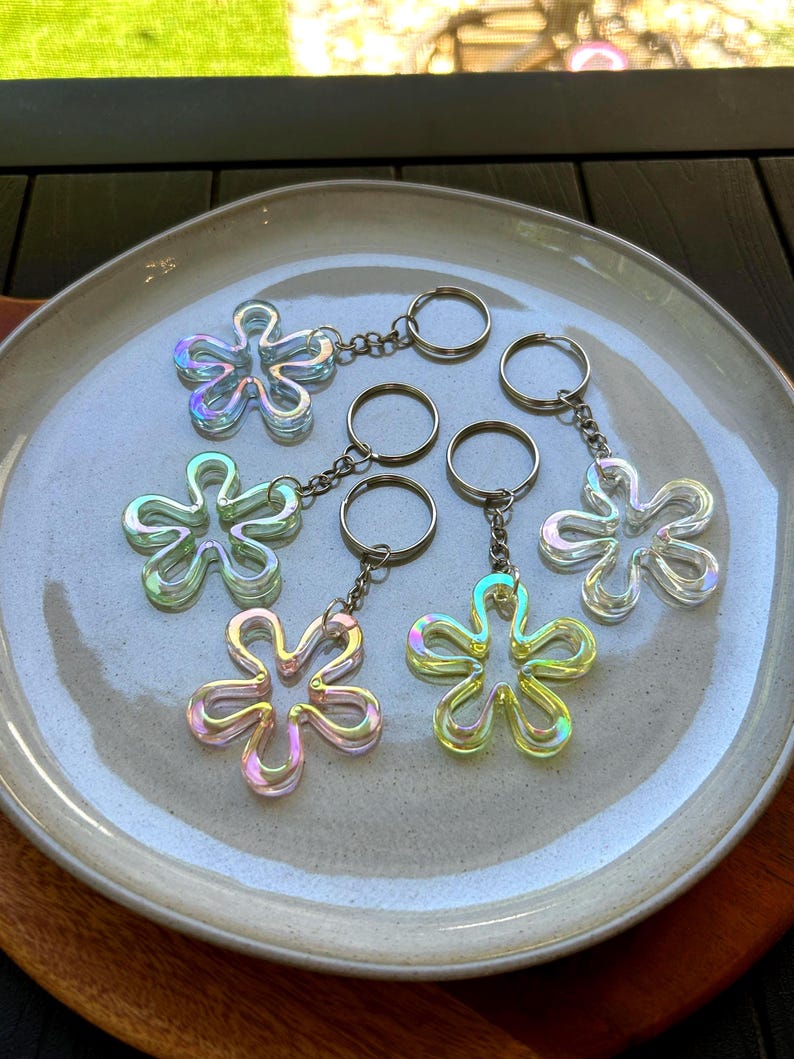 May include: Five flower-shaped keychains with an iridescent, translucent finish. Each keychain features a metal split ring attached by a short chain. The flower charms have a smooth, rounded outline and display shifting colors of light blue, green, and pink.