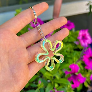 May include: A flower-shaped keychain featuring a translucent, iridescent material that shifts between yellow, green, and pink hues. The charm is attached to a silver-toned metal split ring by a short chain, creating a reflective and glossy accessory.