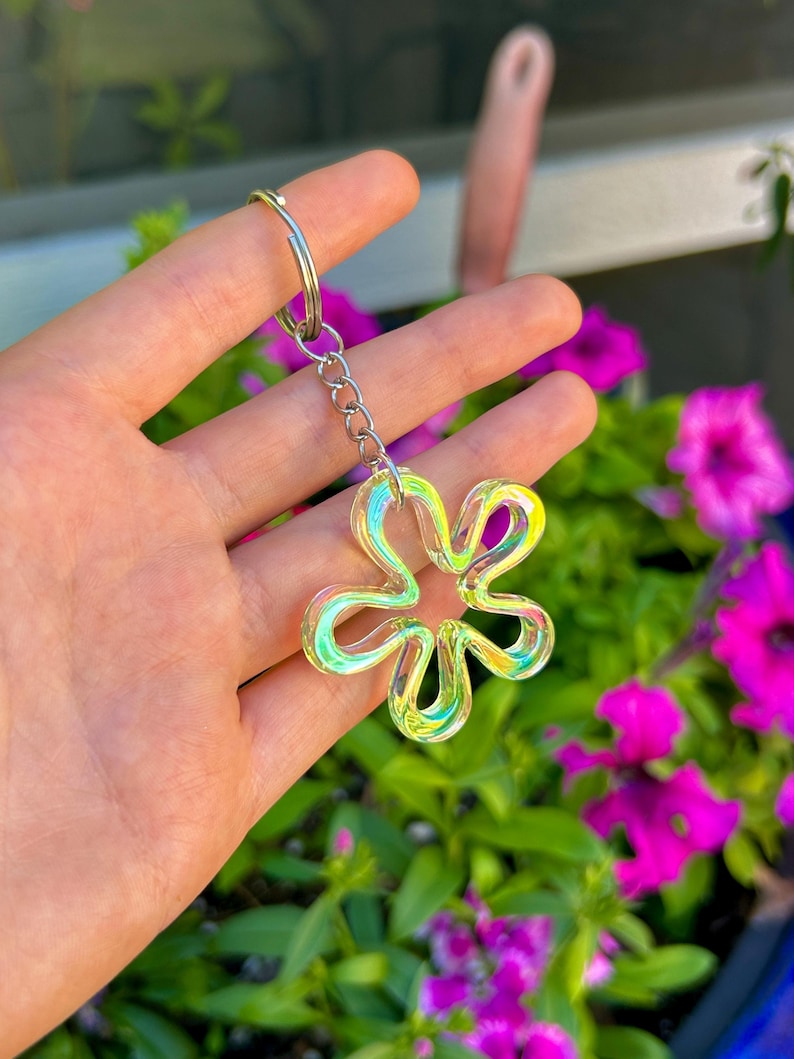 May include: A flower-shaped keychain featuring a translucent, iridescent material that shifts between yellow, green, and pink hues. The charm is attached to a silver-toned metal split ring by a short chain, creating a reflective and glossy accessory.