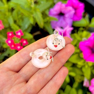 May include: A pair of pale pink glass doughnut-shaped earrings featuring a small white bird figure with a red beak and black eyes attached to the side, suspended from silver-toned metal earring hooks.