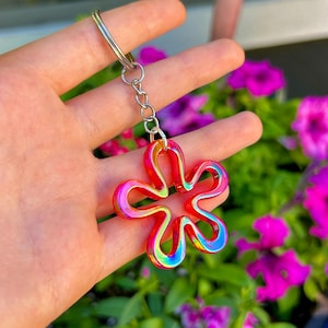 May include: A keychain featuring a five-petaled flower charm with a vibrant, iridescent finish that shifts between shades of red, orange, and blue. The charm is attached to a silver-toned metal split ring by a short, delicate metal chain link.