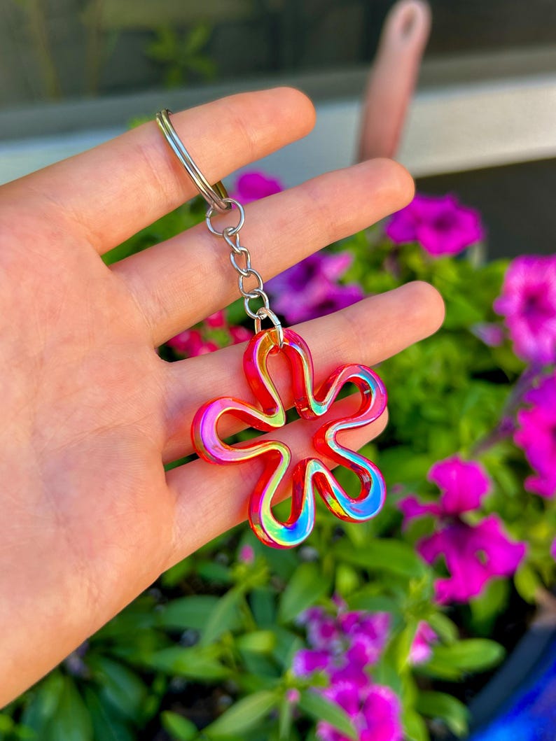 May include: A keychain featuring a five-petaled flower charm with a vibrant, iridescent finish that shifts between shades of red, orange, and blue. The charm is attached to a silver-toned metal split ring by a short, delicate metal chain link.