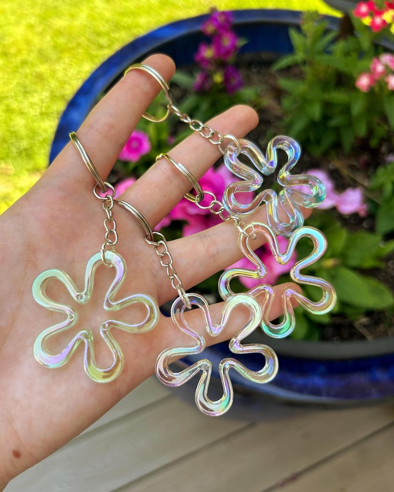 May include: Three iridescent flower-shaped keychains, each featuring a translucent, multi-colored finish that shifts in the light. The charms are attached to silver-toned metal split rings by short, delicate silver-toned metal link chains.