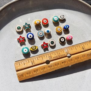 May include: A collection of small, colorful enamel eye-shaped charms and star-shaped charms with gold-colored metal edges. A wooden ruler with inch measurements is placed next to the charms. The charms are arranged on a light gray ceramic plate.