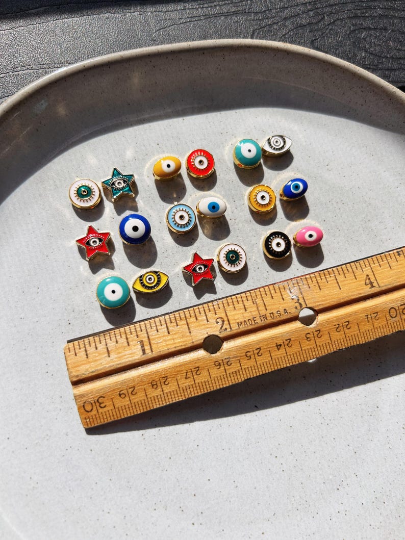 May include: A collection of small, colorful enamel eye-shaped charms and star-shaped charms with gold-colored metal edges. A wooden ruler with inch measurements is placed next to the charms. The charms are arranged on a light gray ceramic plate.