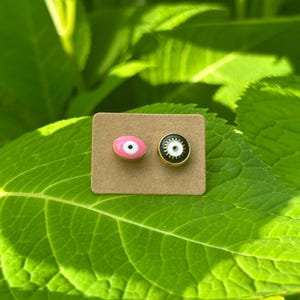 May include: Two stud earrings with an eye design, one pink and one black, each with a gold-colored border. The earrings are on a small, rectangular, brown card, set against a backdrop of green leaves.