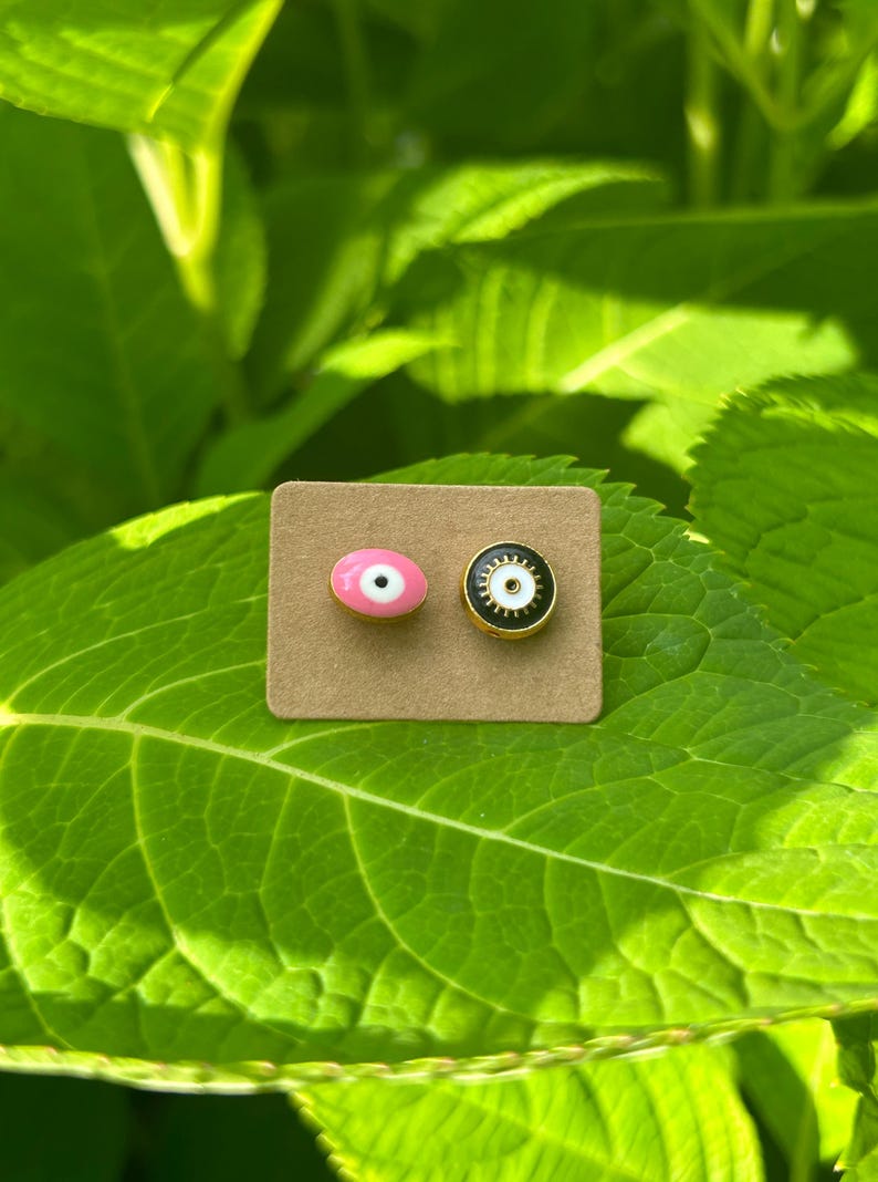 May include: Two stud earrings with an eye design, one pink and one black, each with a gold-colored border. The earrings are on a small, rectangular, brown card, set against a backdrop of green leaves.