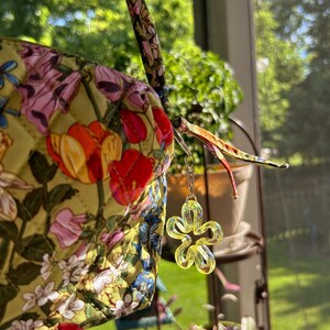 May include: A quilted fabric pouch with a yellow base featuring a colorful floral print of tulips and other blossoms. The pouch includes a matching fabric strap and a silver-toned metal chain charm with a translucent yellow flower pendant.