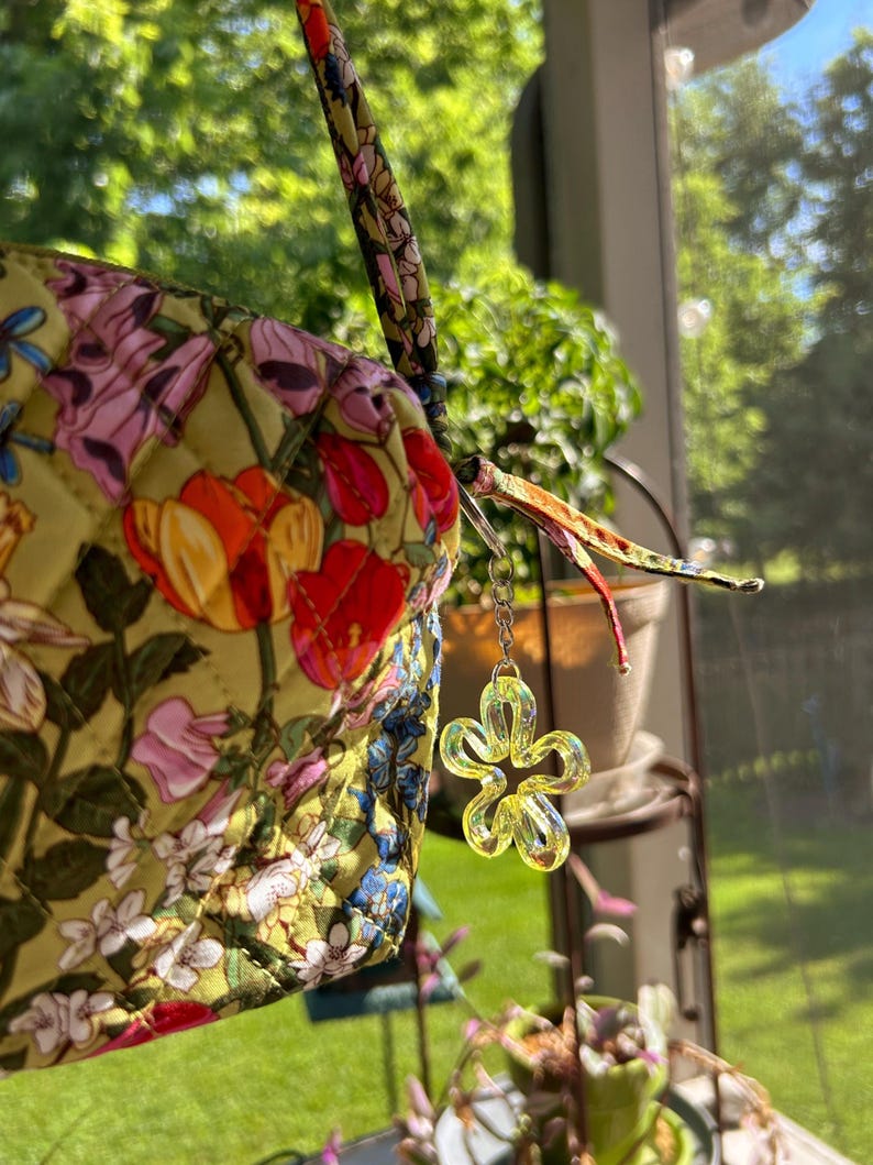 May include: A quilted fabric pouch with a yellow base featuring a colorful floral print of tulips and other blossoms. The pouch includes a matching fabric strap and a silver-toned metal chain charm with a translucent yellow flower pendant.