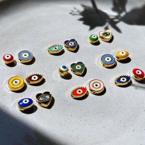 May include: A collection of gold-toned, enamel evil eye charms in various shapes, including round, oval, and heart. The charms feature vibrant colors like blue, red, yellow, and green, with a central eye design. They are displayed on a light gray surface.
