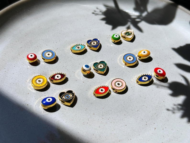 May include: A collection of gold-toned, enamel evil eye charms in various shapes, including round, oval, and heart. The charms feature vibrant colors like blue, red, yellow, and green, with a central eye design. They are displayed on a light gray surface.