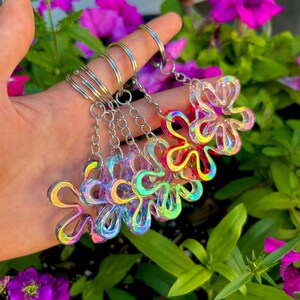 May include: A collection of four flower-shaped keychains made of a translucent, iridescent material that reflects multiple colors. Each keychain features a silver-toned metal split ring attached to a short chain, creating a shimmering effect.