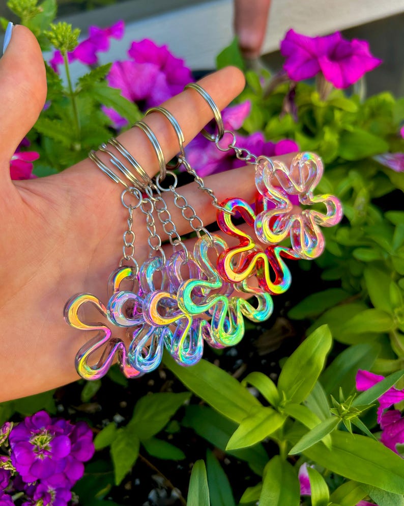 May include: A collection of four flower-shaped keychains made of a translucent, iridescent material that reflects multiple colors. Each keychain features a silver-toned metal split ring attached to a short chain, creating a shimmering effect.