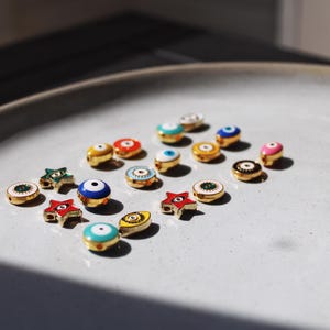 May include: A collection of small, colorful enamel charms with gold-colored metal accents. The charms are in various shapes, including round, star, and eye designs, and are arranged on a light gray plate. The charms feature shades of blue, red, green, and pink.