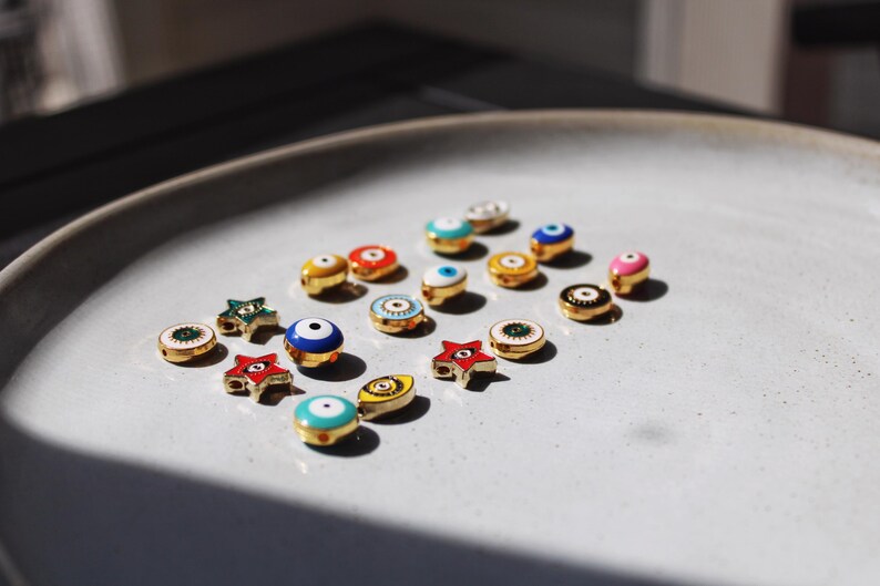 May include: A collection of small, colorful enamel charms with gold-colored metal accents. The charms are in various shapes, including round, star, and eye designs, and are arranged on a light gray plate. The charms feature shades of blue, red, green, and pink.