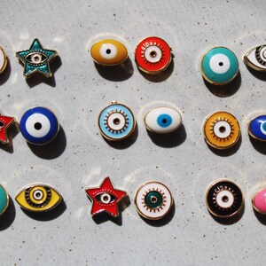 May include: A collection of colorful enamel eye-shaped and star-shaped pins with gold-colored metal trim. The pins feature various colors, including red, blue, yellow, green, and pink, with a white or black pupil design. The pins are arranged on a light gray surface.