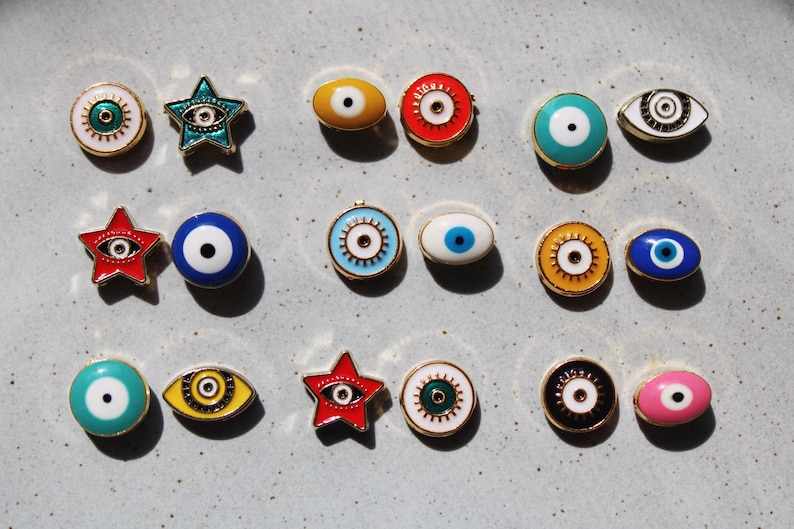 May include: A collection of colorful enamel eye-shaped and star-shaped pins with gold-colored metal trim. The pins feature various colors, including red, blue, yellow, green, and pink, with a white or black pupil design. The pins are arranged on a light gray surface.