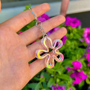 May include: A flower-shaped keychain featuring a translucent, iridescent material that reflects multiple colors. The charm is attached to a silver-toned metal split ring by a short chain, creating a reflective and glossy appearance in the light.