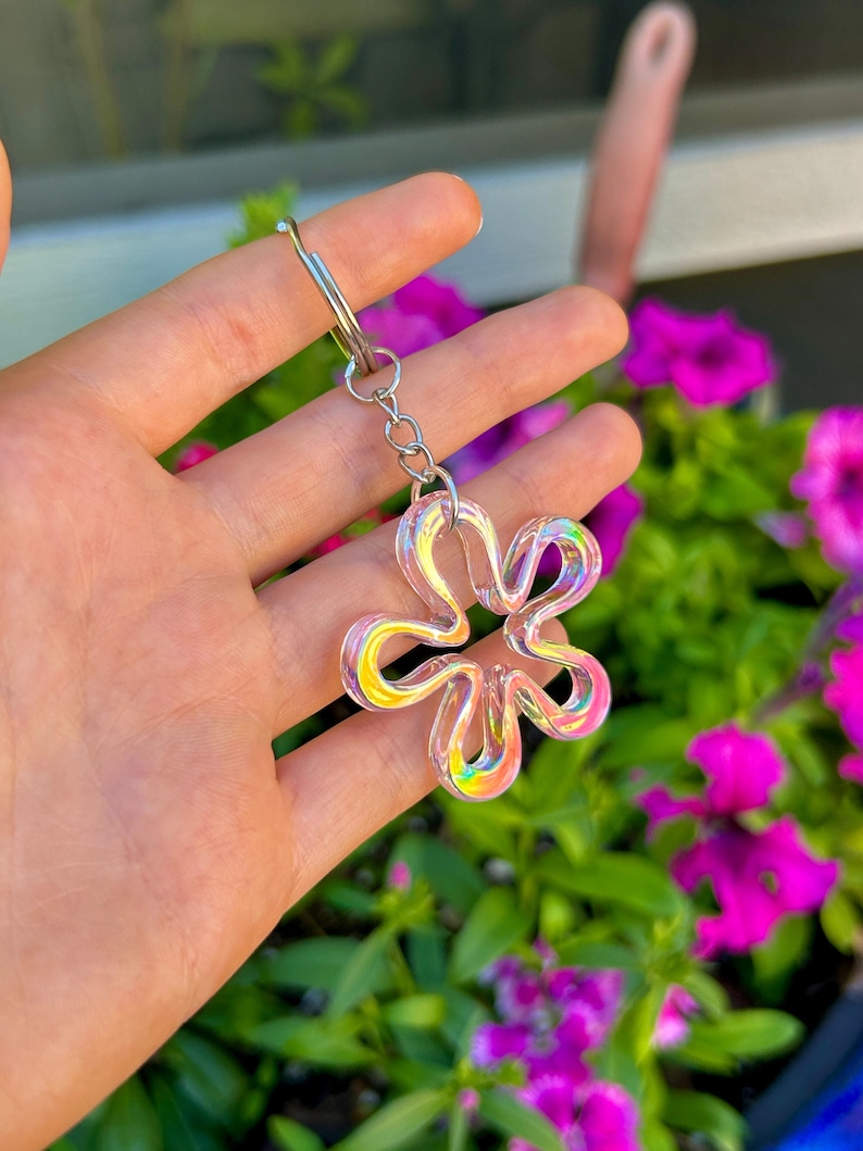 May include: A flower-shaped keychain featuring a translucent, iridescent material that reflects multiple colors. The charm is attached to a silver-toned metal split ring by a short chain, creating a reflective and glossy appearance in the light.