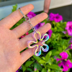 May include: A flower-shaped keychain featuring a translucent, iridescent material that shifts between shades of blue, purple, and yellow. The charm is attached to a silver-toned metal split ring by a short, delicate silver-toned metal chain link.