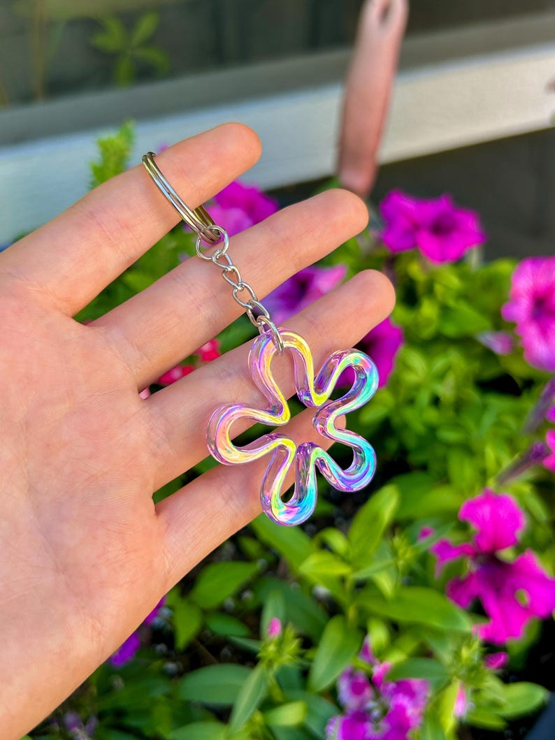 May include: A flower-shaped keychain featuring a translucent, iridescent material that shifts between shades of blue, purple, and yellow. The charm is attached to a silver-toned metal split ring by a short, delicate silver-toned metal chain link.