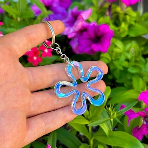 May include: A translucent, iridescent flower-shaped keychain with a smooth, rounded edge design. The charm features a shifting color palette of blue, purple, and white tones, attached to a standard silver-toned metal split ring and chain.
