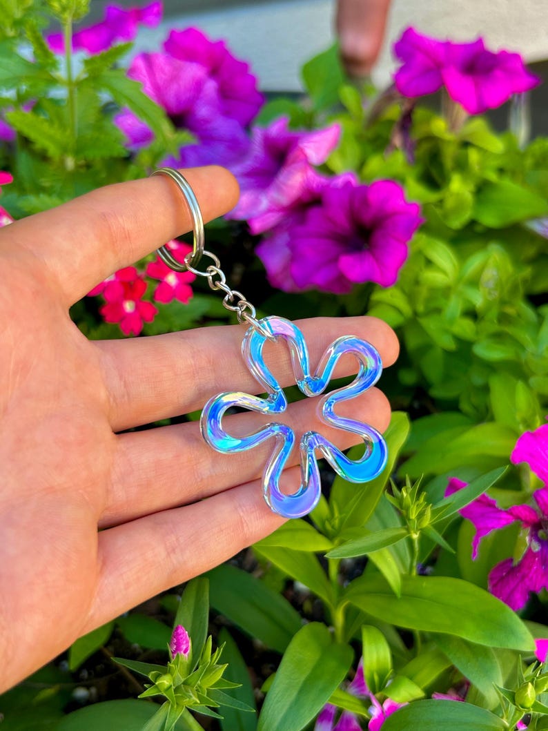 May include: A translucent, iridescent flower-shaped keychain with a smooth, rounded edge design. The charm features a shifting color palette of blue, purple, and white tones, attached to a standard silver-toned metal split ring and chain.