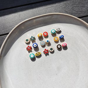 May include: A collection of colorful enamel charms, including eye and star designs, arranged on a light gray ceramic plate. The charms feature various colors like blue, red, yellow, and green, with black and white detailing. The plate sits on a dark wood surface.