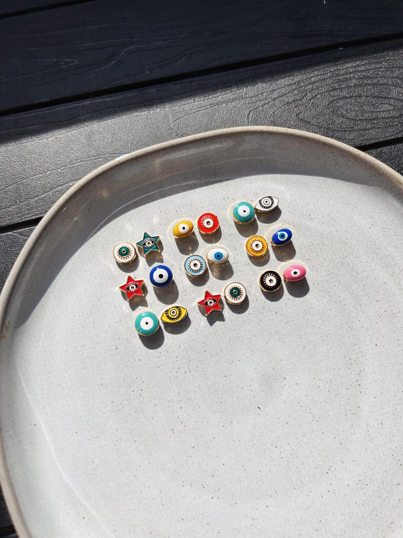 May include: A collection of colorful enamel charms, including eye and star designs, arranged on a light gray ceramic plate. The charms feature various colors like blue, red, yellow, and green, with black and white detailing. The plate sits on a dark wood surface.
