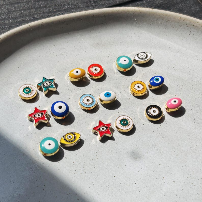 May include: A collection of colorful enamel evil eye charms in various shapes, including round, oval, and star. The charms feature gold-colored metal accents and come in a variety of colors, including blue, red, yellow, and green.